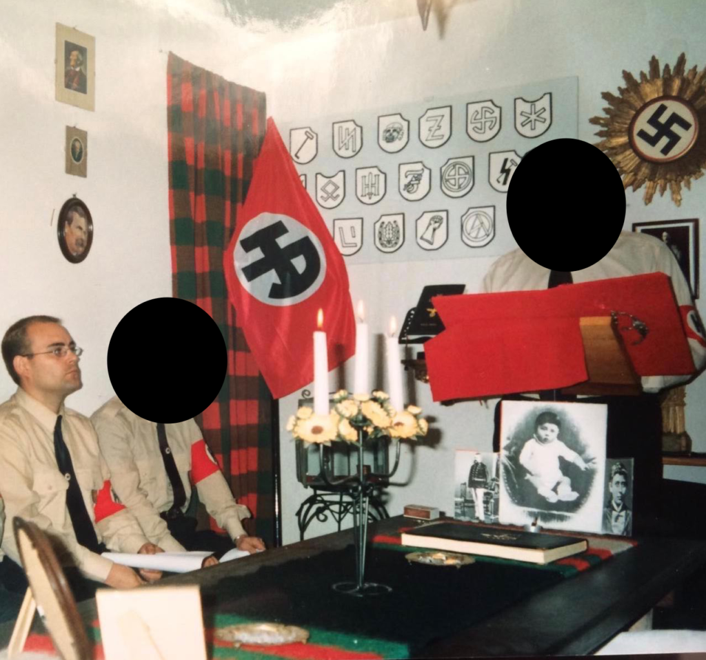 Jorge Bonito participando en una ceremonia pagana nazi. En la mesa, los retratos de Adolf Hitler de niño y de sus padres, Klaus y Alois. Junto al resto de símbolos nazis, aparece la bandera de Hermandad Aria con la H y la A ('Hereditas et ambitus') entrelazadas simulando la esvástica nazi. 